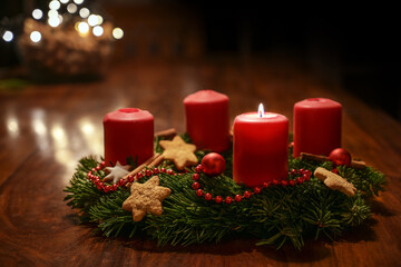 First Advent - decorated Advent wreath from fir branches with red burning candles on a wooden table in the time before Christmas, festive bokeh in the warm dark background, copy space, selected focus - obrazy, fototapety, plakaty