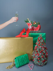 Hand with a glass of shompansky and beautifully wrapped gifts and Christmas tree decorations on a gray background. There is room for text. 
