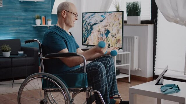 Pensioner sitting in wheelchair and doing exercise with dumbbells while looking at video of training lesson on tablet. Man with physical disability watching workout video, lifting weights
