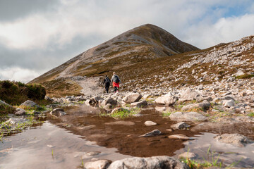Fototapeta na wymiar Couple of women walking on a path to the top of Croagh Patrick mountain. County Mayo, Ireland. Popular landmark for pilgrimage and hiking. Clean cloudy sky. Peak reflection in a puddle of water.
