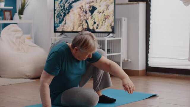 Pensioner sitting on yoga mat to meditate after training. Senior woman in lotus position doing meditation as zen relaxation. Elder person meditating for wellness and mindfulness.