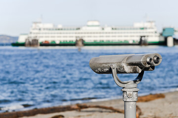 A pair of metal binoculars on a fixed stand look out over an ocean and a ferry boat which are out...