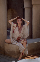 red-haired beautiful girl in Arabic clothes sit against the background of the old arabic city, bazaar. dressed in a beige abaya with embroidery and a scarf on her head