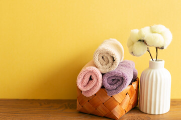 Obraz na płótnie Canvas Stack of bath towels in basket and cotton plant on wooden table. yellow background. copy space