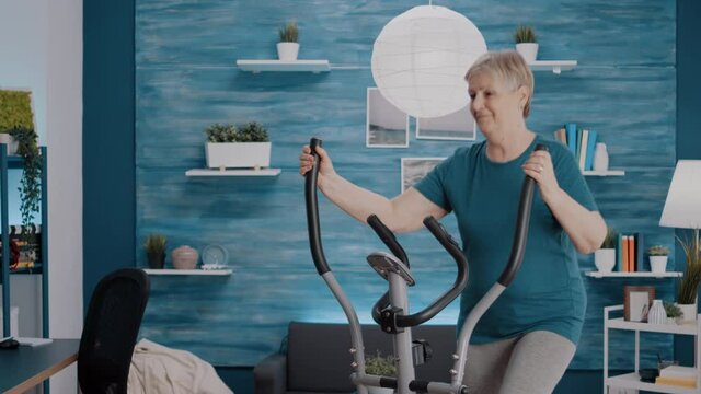 Mature woman using stationary bicycle to do physical exercise at home. Retired person training legs muscles with electronic stepper bike, doing fitness workout with cardio machine.
