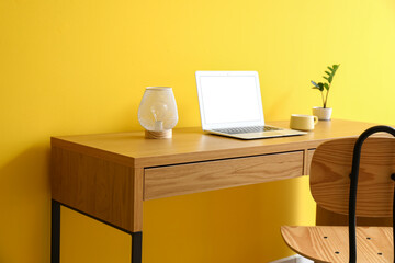 Wooden table with modern laptop, lamp, cup and houseplant near yellow wall