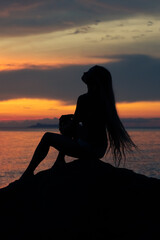 Silhouette of a young woman with a long hair sitting on the rock by the sea and enjoying colorful sunset