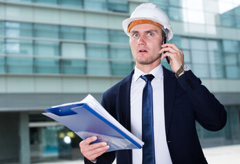 Architector in suit and hat is taking folder and considering project by the phone near the building.