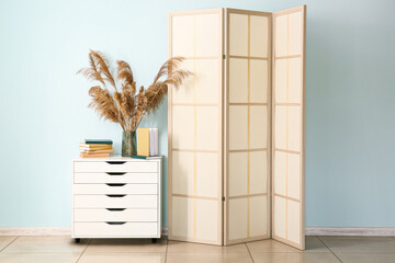 Chest of drawers with vase, books and folding screen near blue wall