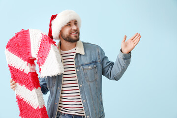 Handsome man in Santa hat with candy cane pinata showing something on blue background