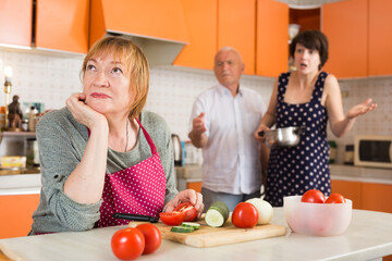 Upset elderly woman in apron sitting at kitchen table and cutting vegetables while her husband and adult daughter reprimanding her