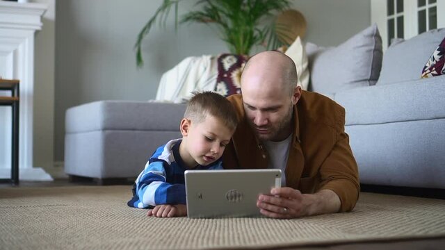 Father spends time with little son. Positive young man with cute little child play videogame on tablet computer lying on floor near sofa at home spbd