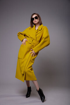 High fashion photo of a beautiful elegant young woman in a pretty yellow mustard coat, black ankle boots, stylish sunglasses posing over gray background. Slim figure. Bob haircut. Studio Shot.