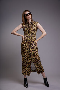 High fashion photo of a beautiful elegant young woman in a pretty shirt dress sleeveless animal leopard print, black ankle boots, stylish sunglasses posing over gray background. Slim figure.