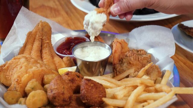 Woman's hand picks up shrimp from mixed fried seafood platter and dips into tartar sauce, slider 4K