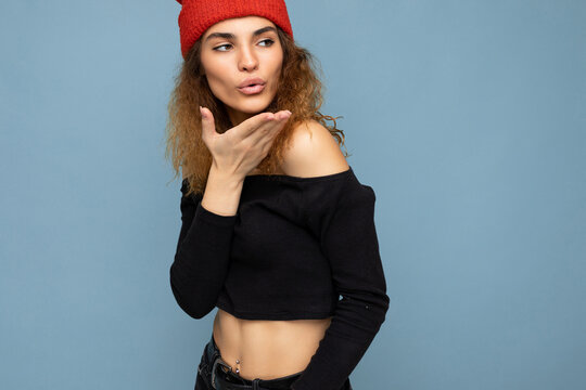 Photo of young positive cute nice brunette woman curly with sincere emotions wearing stylish black crop top and red hat isolated on blue background with copy space and giving kiss