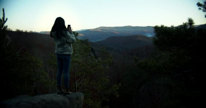 Wide angle of asian woman traveler taking photos with mobile phone, mountains and forest on the background. Female traveler standing with beautiful landscape on the sunset. Travelling concept.