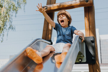 cheerful little boy in a blue T-shirt and white shorts rolls down a metal slide in city park. child...