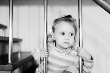 A little girl peeks out of the stairs through the railing. Black and white photo. Front view.