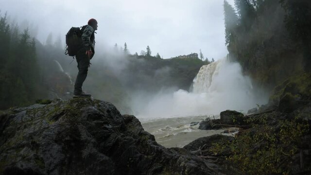 Hiker with Backpack Looking Up at Massive Waterfall in Extreme Conditions