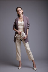 High fashion photo of a beautiful elegant young woman in a pretty purple jacket, white top, pants, accessories, handbag posing on gray background. Studio Shot. Slim figure. Make up, hairstyle