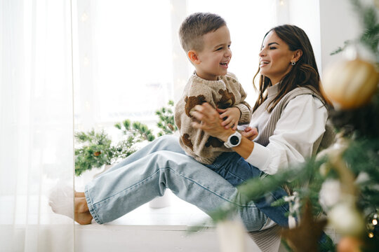 Happy mother with son sit on windowsill near Christmas tree