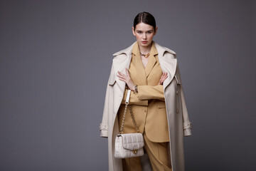 High fashion photo of a beautiful elegant young woman in a pretty white coat, beige sand suit, jacket, pants, trousers, accessories, handbag posing on gray background. Studio Shot. Slim figure.