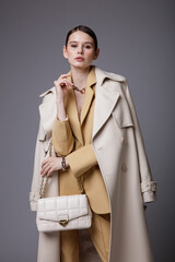 High fashion photo of a beautiful elegant young woman in a pretty white coat, beige sand suit, jacket, pants, trousers, accessories, handbag posing on gray background. Studio Shot. Slim figure.