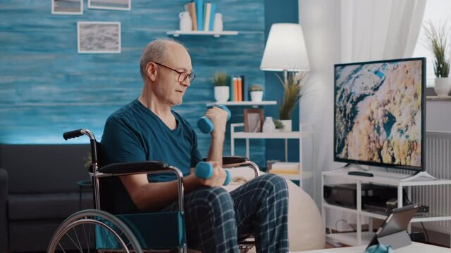 Retired man sitting in wheelchair and lifting dumbbells to exercise while watching online video of workout on tablet. Elder adult with disability using weights, following training lesson