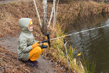 The child is a fisherman by the autumn lake. A child in warm clothes in the rain sits on the shore with a fishing rod