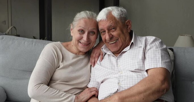 Happy older adult spouses sit on couch hug with warmth look at camera with healthy white smiles pose for family photo portrait together. Beautiful positive aged loving couple cuddle on sofa at home