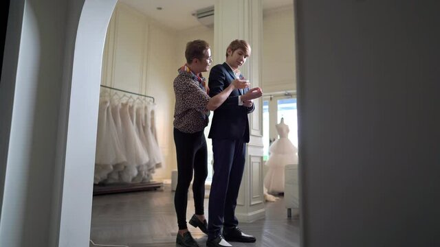 4K Asian LGBTQ guy bridal shop owner helping male groom customer choosing and trying on wedding suit at wedding studio. Small business entrepreneur wedding planner and marriage ceremony concept