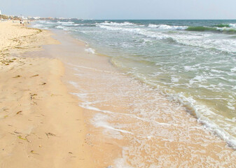 Sandy beach coast seascape. Unidentified people on background. Smooth foamy waves crash on seashore...