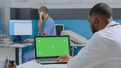 African american doctor looking at mock up green screen chroma key laptop computer with isolated display. Therapist man analyzing sickness diagnostic working at medical report in hospital office