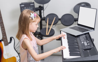 Home lesson of music. Cute little blonde girl learning to play piano keyboard synthesizer watching online lesson on laptop, drum set and guitar on blurred background. Studying the music at home