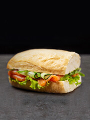 Italian Ciabatta bread sandwich with raw salmon and cucumber slices, fresh lettuce and arugula served without plate on dark concrete background. Copy space text image
