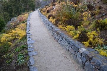 road cut into the slope. above and below the road is a stone dry wall. nature trail through the autumn park with a drain and a metal grid. slopes overgrown with drought perennials yellow, yew, boulder