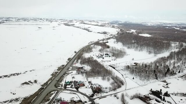 Aerial shot in movement of snowed village in right and field in left with road between and distant forest. Winter or early spring