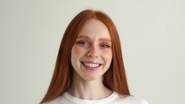Close-up face of charming smiling young woman touching playfully red hair looking at camera, on white isolated background in studio. Happy atractive lady flirting with you, slow motion.