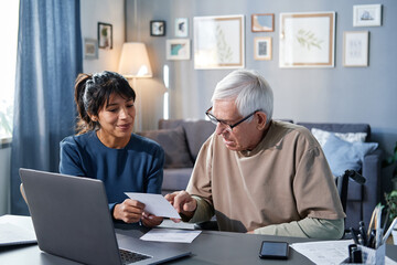 Senior man pointing at bill while sitting at the table with laptop and woman who helping him in online payment at home