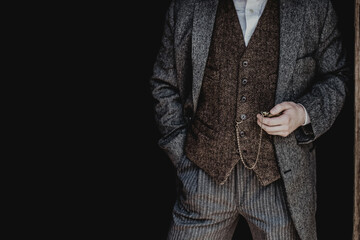A man in a gray tweed suit and a brown vest holds a retro watch on a chain. Men's clothing banner...