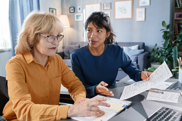 Obraz premium Senior businesswoman in eyeglasses working on laptop with documents at the table together with assistant, they working at home