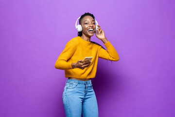 African American woman wearing headphones listening to music from smart phone on colorful purple isolated studio background