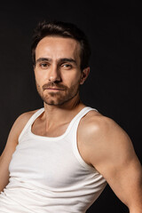 Fototapeta premium Close-up portrait of young handsome bearded man in white undershirt isolated on dark studio background. Emotions, facial expression