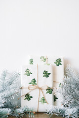 the New Year's gift is packed in festive paper with watercolor Christmas trees on a wooden table against a white wall with white spruce branches