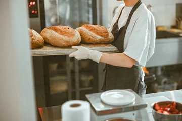 Poster Woman holds tray with large breads standing in spacious craft bakery © Friends Stock