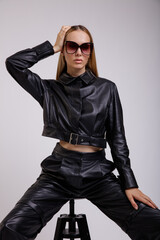 High fashion photo of a beautiful elegant young woman in a pretty leather suit, pants, jacket, stylish sunglasses posing on white background. Studio Shot. Slim figure. Model is sitting on chair