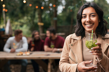 young indian casual woman with attractive smile holding a cocktail in hand in the foreground - friends sitting at table defocused background