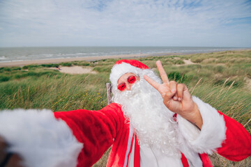 happy santa claus on holiday at the sea making victory sign on phone camera - selfie and holiday...