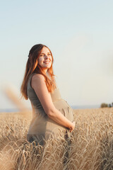 beautiful young pregnant woman walks on wheat field at sunset, expectant mother with relax in nature stroking her belly with hand, happy pregnancy concept